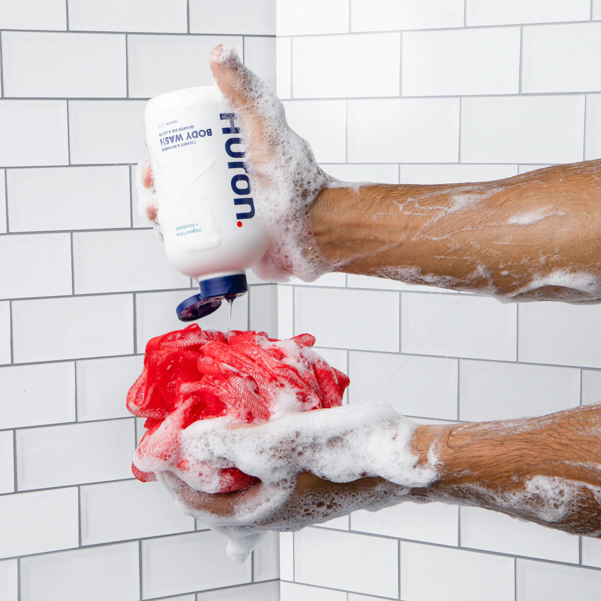 Body wash gets squeezed onto a shower loofah in a white tiled shower.