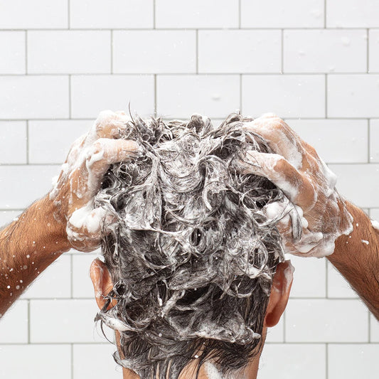A man washes his hair in a white tiled shower. 