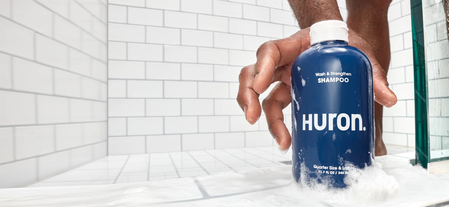 A hand reaches for a bottle of shampoo in a white tiled shower. 