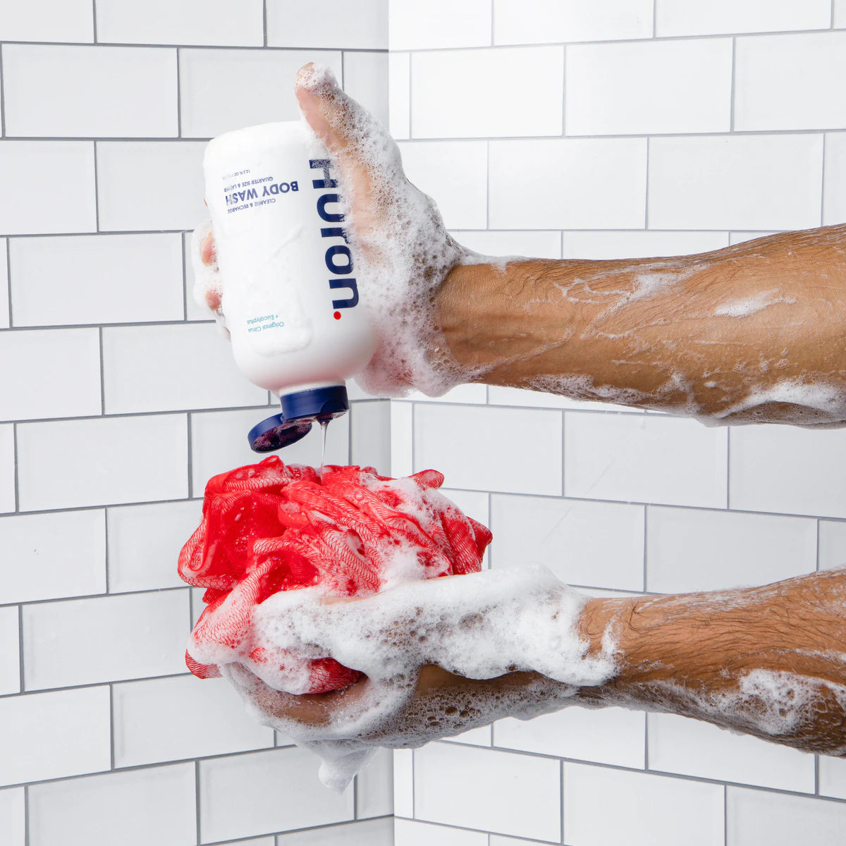 Body wash gets squeezed onto a shower loofah in a white tiled shower. 