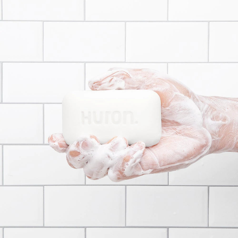 A hand holds a lathery bar of soap against a white tiled background. 